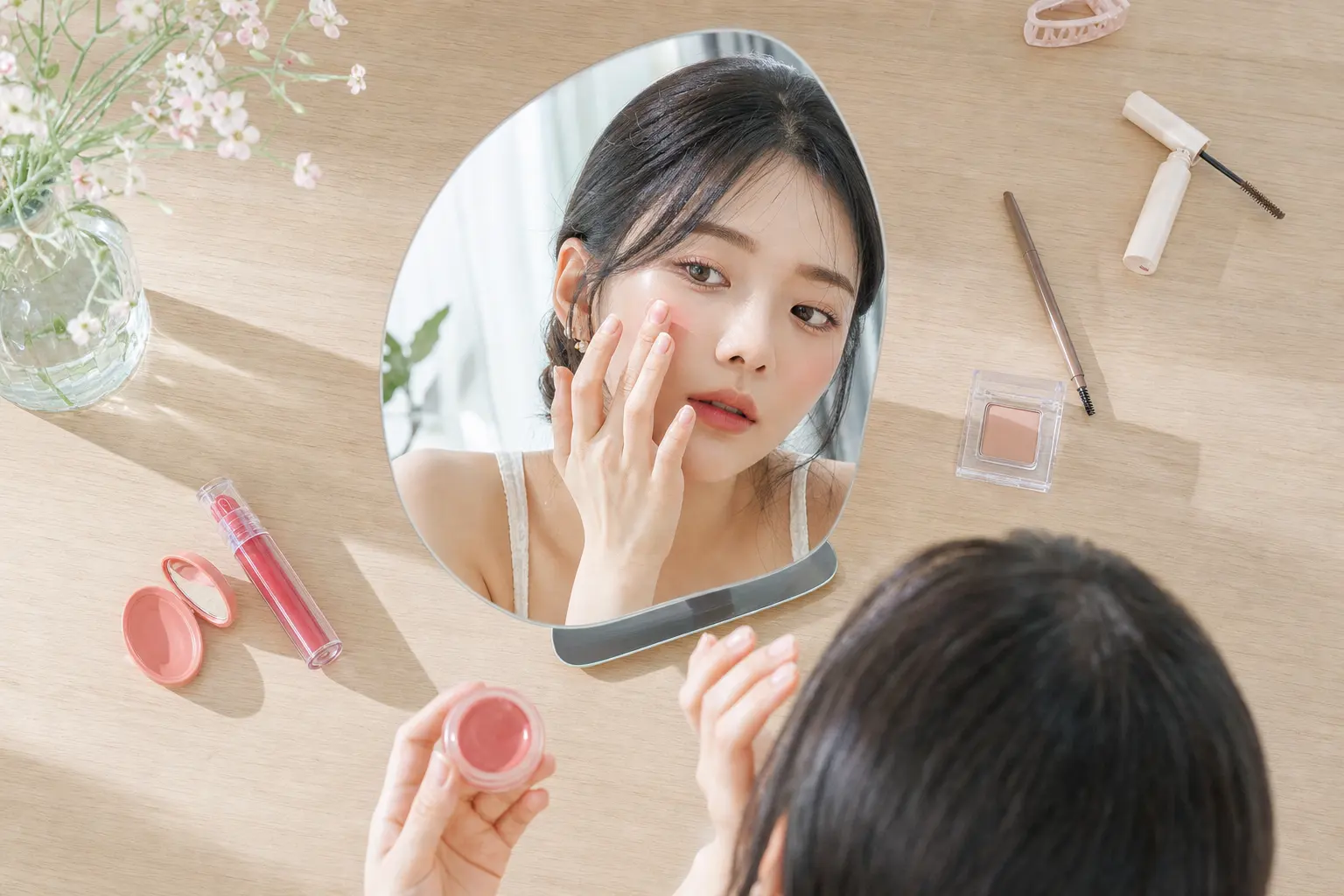 Overhead view of a young woman’s face and hands at a desk mirror as she applies cream blush with her fingers, surrounded