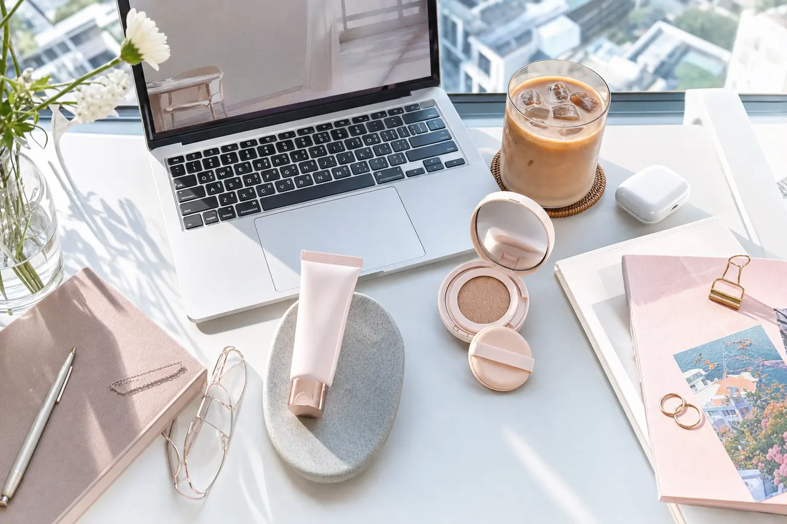 Desk setup with a laptop, iced coffee, and a Korean sunscreen next to an SPF cushion compact, showing a mid-day reapplic