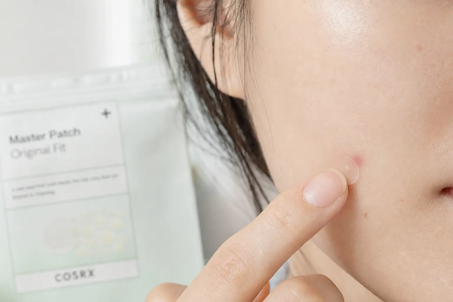 Close crop of a finger applying a clear hydrocolloid patch to a small whitehead on a cheek; natural skin texture visible