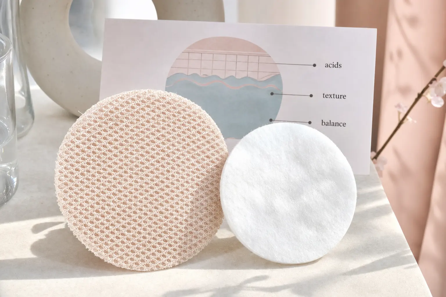 Create a close-up educational still life of a dense mesh facial pad beside a simple cotton pad, with a small abstract di